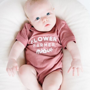 May include: A baby wearing a dusty rose onesie with the text "FLOWER FARMER" and a small floral design. The baby is lying on a white surface.