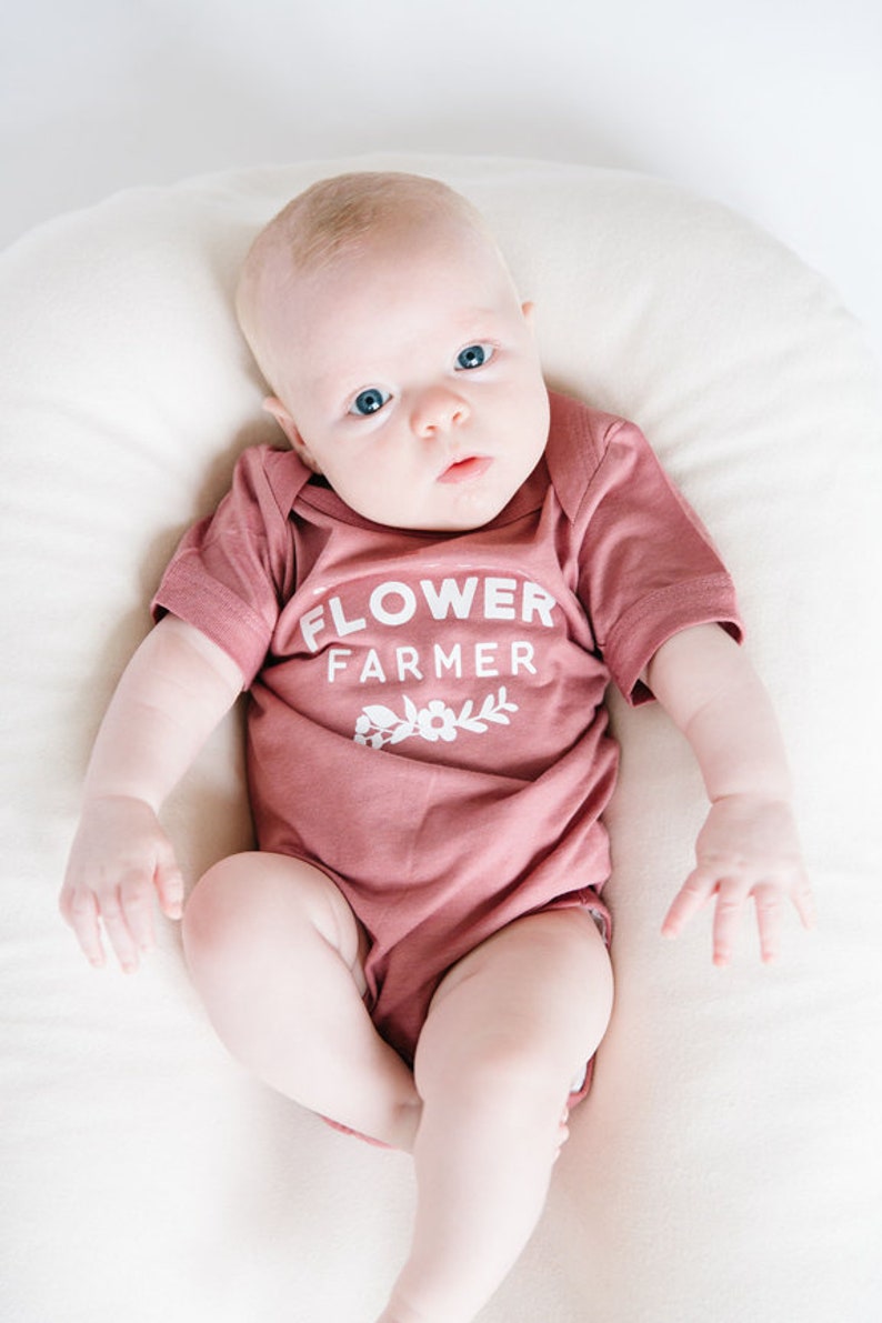 May include: A baby wearing a dusty rose onesie with the text "FLOWER FARMER" and a small floral design. The baby is lying on a white surface.