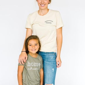 May include: A woman and a child wearing matching t-shirts. The woman's shirt says "Homeschool Mama" in black text. The child's shirt says "home schooled" in white text. The woman is wearing blue jeans and the child is wearing khaki pants.