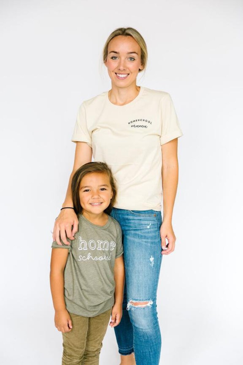 May include: A woman and a child wearing matching t-shirts. The woman's shirt says "Homeschool Mama" in black text. The child's shirt says "home schooled" in white text. The woman is wearing blue jeans and the child is wearing khaki pants.
