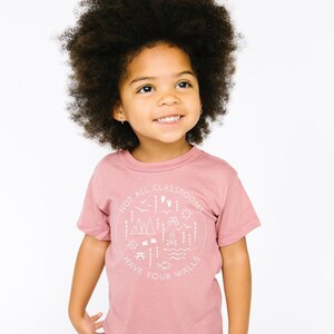 Puede incluir: Una persona joven lleva una camiseta rosa con un gr&aacute;fico blanco que dice "No todas las aulas tienen cuatro paredes". El gr&aacute;fico incluye ilustraciones de monta&ntilde;as, &aacute;rboles, una fogata, una br&uacute;jula, un pez y otros elementos al aire libre. La persona lleva pantalones vaqueros azules y tiene el pelo rizado.