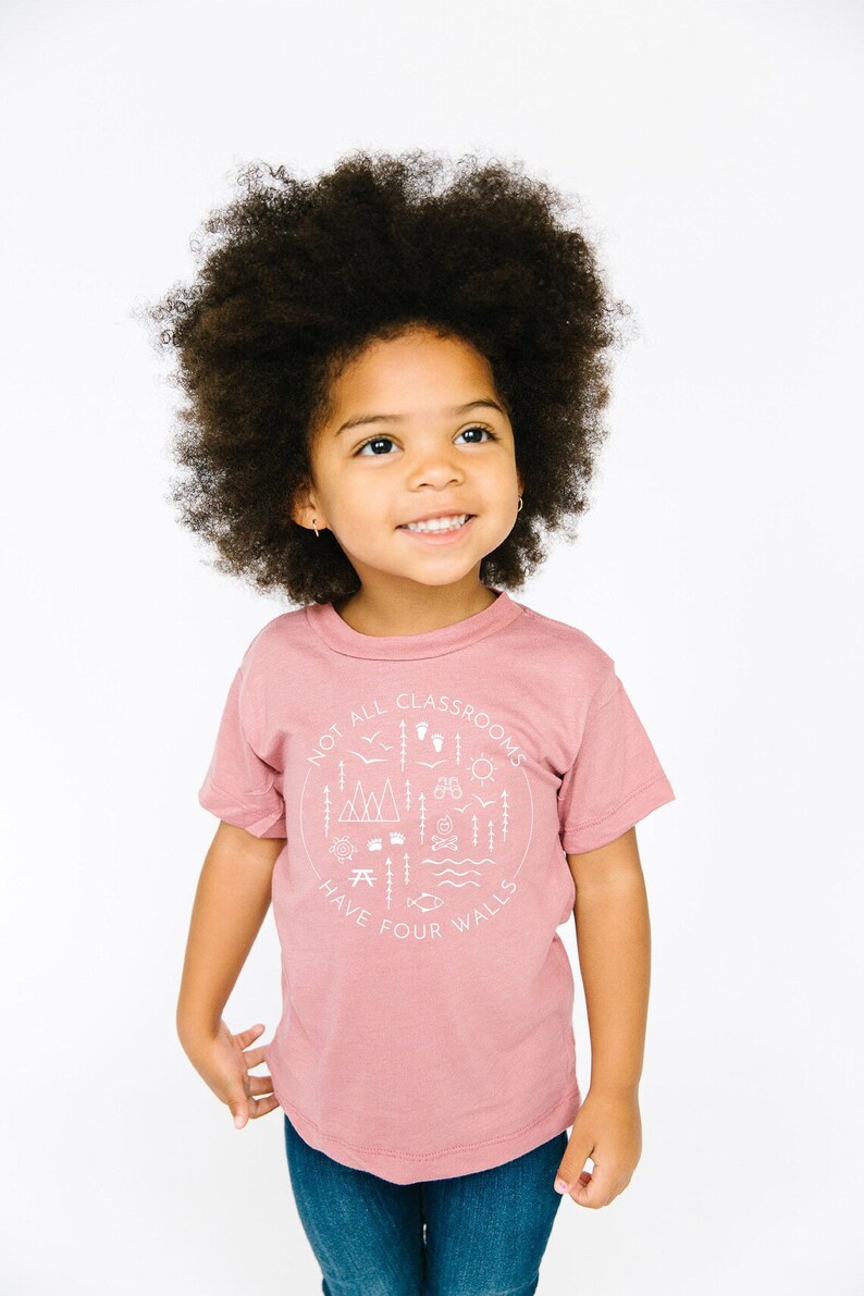 Puede incluir: Una persona joven lleva una camiseta rosa con un gr&aacute;fico blanco que dice "No todas las aulas tienen cuatro paredes". El gr&aacute;fico incluye ilustraciones de monta&ntilde;as, &aacute;rboles, una fogata, una br&uacute;jula, un pez y otros elementos al aire libre. La persona lleva pantalones vaqueros azules y tiene el pelo rizado.