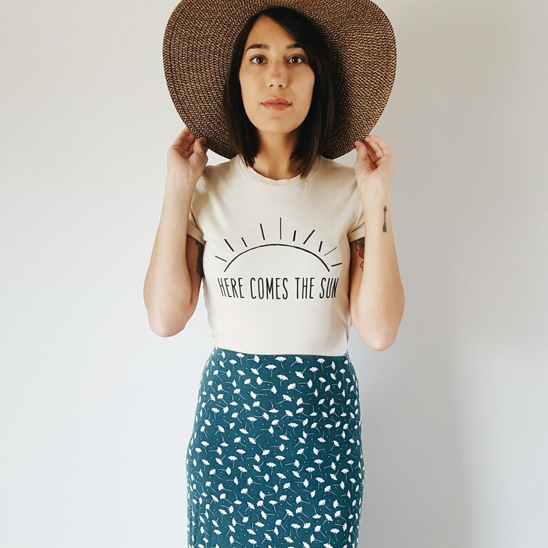 K&ouml;nnte beinhalten: Eine Frau tr&auml;gt ein cremefarbenes T-Shirt mit dem Text "Here Comes The Sun" und einer Sonnengrafik. Sie tr&auml;gt au&szlig;erdem einen breitkrempigen braunen Hut und einen blaugr&uuml;nen Rock mit wei&szlig;em Blumenmuster.