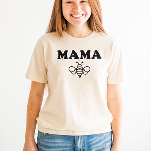 K&ouml;nnte beinhalten: Ein cremefarbenes T-Shirt mit einem schwarzen Aufdruck, der "MAMA" und ein Bienendesign zeigt.
