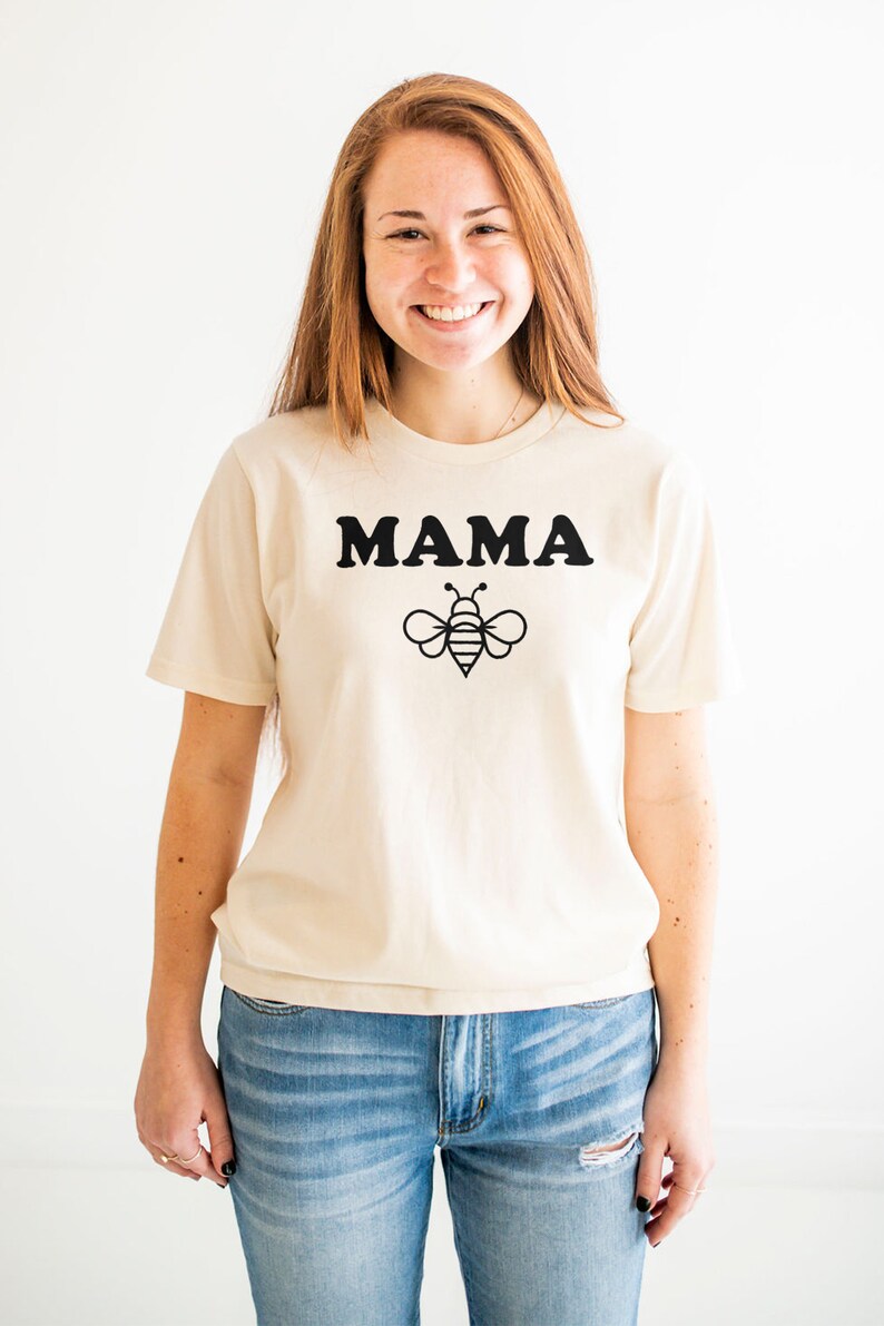 K&ouml;nnte beinhalten: Ein cremefarbenes T-Shirt mit einem schwarzen Aufdruck, der "MAMA" und ein Bienendesign zeigt.