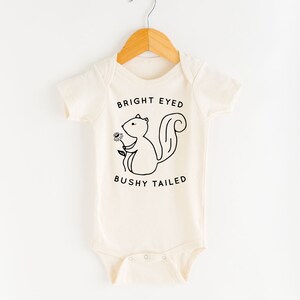 May include: A cream-colored baby onesie with short sleeves features a black and white illustration of a squirrel holding a flower. The text "BRIGHT EYED BUSHY TAILED" is printed below the squirrel.