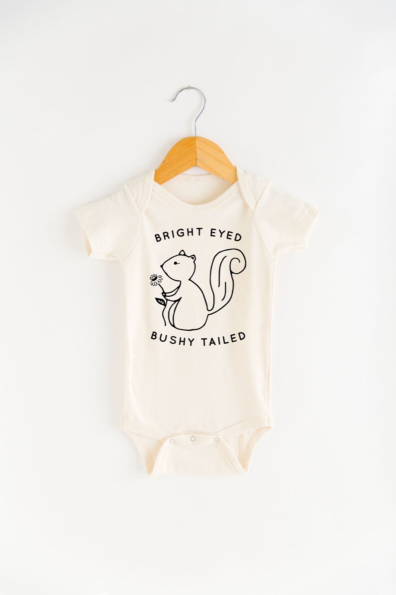 May include: A cream-colored baby onesie with short sleeves features a black and white illustration of a squirrel holding a flower. The text "BRIGHT EYED BUSHY TAILED" is printed below the squirrel.