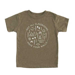 Puede incluir: Una camiseta verde de manga corta con un dise&ntilde;o gr&aacute;fico blanco que dice "Not all classrooms have four walls". El dise&ntilde;o gr&aacute;fico incluye un c&iacute;rculo con varios iconos de exteriores, como monta&ntilde;as, &aacute;rboles, una fogata, una br&uacute;jula y un pez.