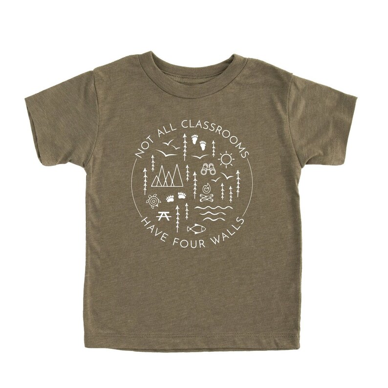 Puede incluir: Una camiseta verde de manga corta con un dise&ntilde;o gr&aacute;fico blanco que dice "Not all classrooms have four walls". El dise&ntilde;o gr&aacute;fico incluye un c&iacute;rculo con varios iconos de exteriores, como monta&ntilde;as, &aacute;rboles, una fogata, una br&uacute;jula y un pez.
