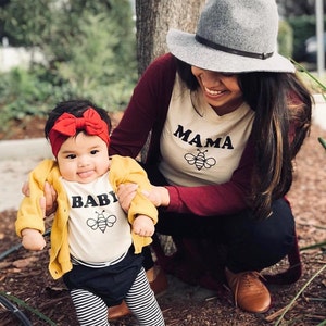 K&ouml;nnte beinhalten: Eine Mutter und ihre Tochter tragen passende Shirts. Das Shirt der Mutter sagt "Mama" mit einer Bienengrafik. Das Shirt der Tochter sagt "Baby" mit einer Bienengrafik. Die Tochter tr&auml;gt ein rotes Stirnband und eine gelbe Strickjacke.