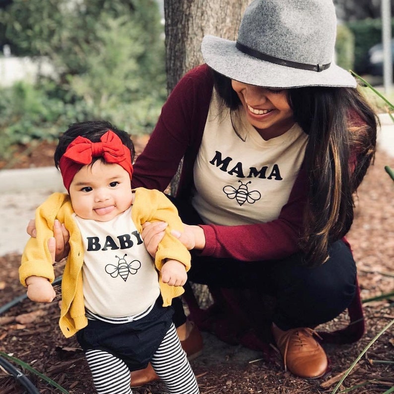 K&ouml;nnte beinhalten: Eine Mutter und ihre Tochter tragen passende Shirts. Das Shirt der Mutter sagt "Mama" mit einer Bienengrafik. Das Shirt der Tochter sagt "Baby" mit einer Bienengrafik. Die Tochter tr&auml;gt ein rotes Stirnband und eine gelbe Strickjacke.