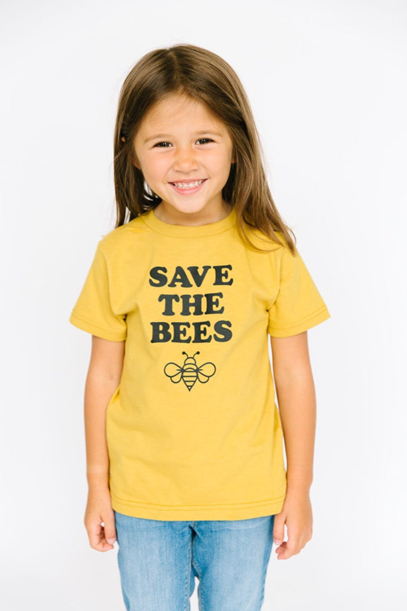 Puede incluir: Una persona joven con una camiseta amarilla con el texto "SAVE THE BEES" y un gr&aacute;fico de abeja negro y amarillo. La persona lleva pantalones vaqueros azules.