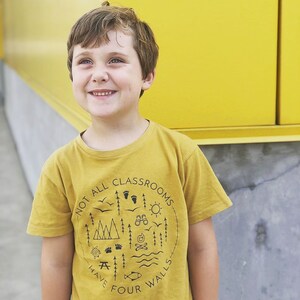 Puede incluir: Un ni&ntilde;o peque&ntilde;o con una camiseta amarilla mostaza con un estampado gr&aacute;fico negro que dice "No todas las aulas tienen cuatro paredes". El gr&aacute;fico incluye ilustraciones de &aacute;rboles, monta&ntilde;as, una fogata, una tienda de campa&ntilde;a, un pez y una mesa de picnic.