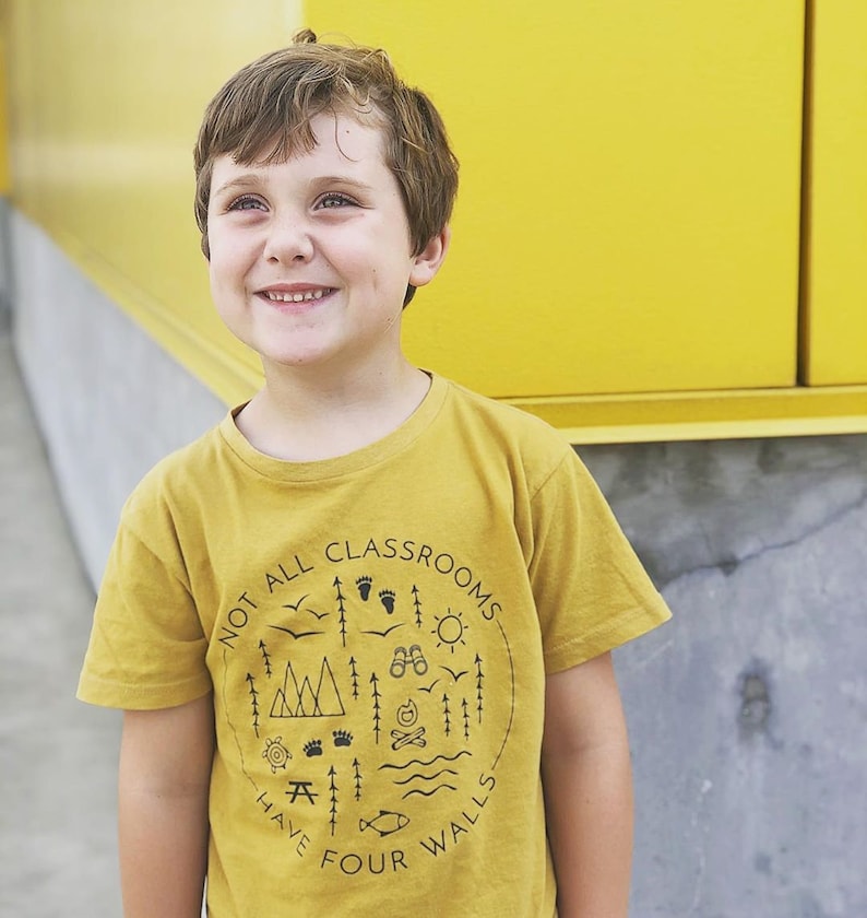 Puede incluir: Un ni&ntilde;o peque&ntilde;o con una camiseta amarilla mostaza con un estampado gr&aacute;fico negro que dice "No todas las aulas tienen cuatro paredes". El gr&aacute;fico incluye ilustraciones de &aacute;rboles, monta&ntilde;as, una fogata, una tienda de campa&ntilde;a, un pez y una mesa de picnic.