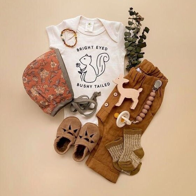 May include: A baby outfit set with a white bodysuit featuring a squirrel graphic and the text "Bright Eyed Bushy Tailed", brown pants, a floral bonnet, brown leather shoes, a wooden deer toy, and a wooden bead teether.