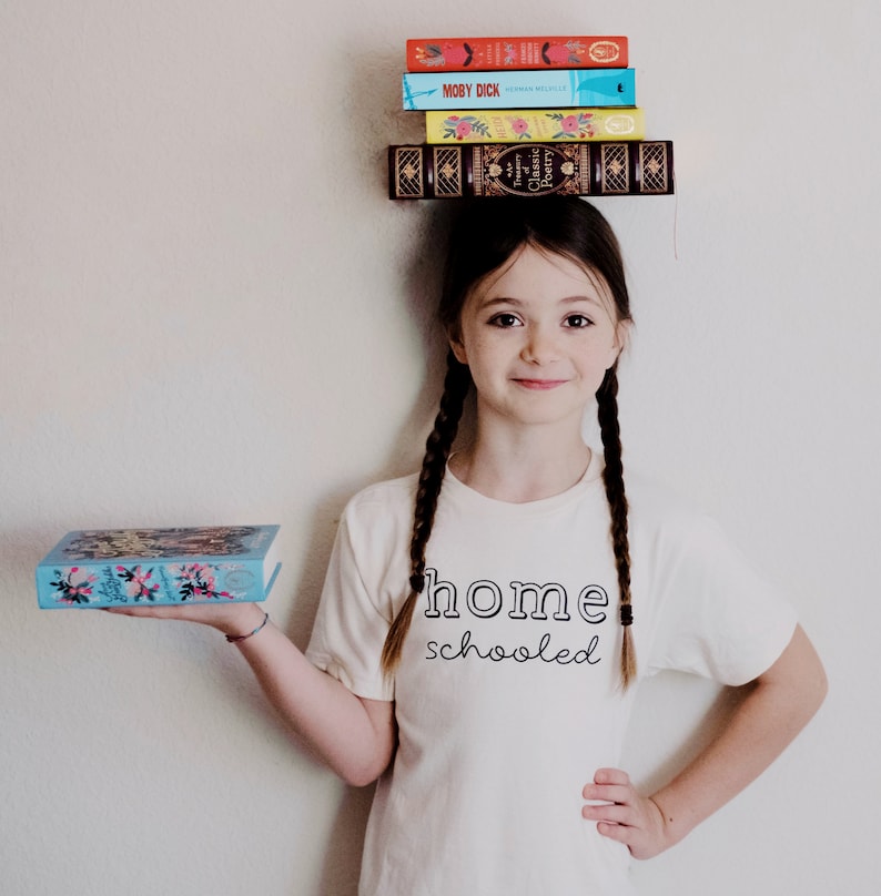 May include: A young girl with long brown braids is wearing a white t-shirt that says "home schooled". She is holding a blue book with colorful flowers on the cover and has a stack of books balanced on her head. The books on her head are a mix of colors and have titles like "Moby Dick" and "Classic Poetry".