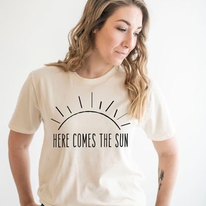 K&ouml;nnte beinhalten: Eine Frau tr&auml;gt ein cremefarbenes T-Shirt mit einer schwarzen Grafik einer Sonne und dem Text "Here Comes The Sun". Sie tr&auml;gt eine blaue Jeans.
