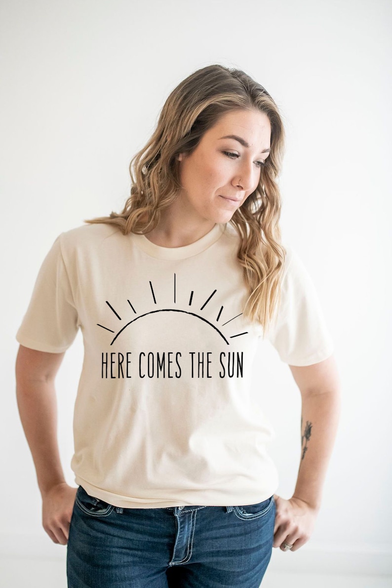 K&ouml;nnte beinhalten: Eine Frau tr&auml;gt ein cremefarbenes T-Shirt mit einer schwarzen Grafik einer Sonne und dem Text "Here Comes The Sun". Sie tr&auml;gt eine blaue Jeans.