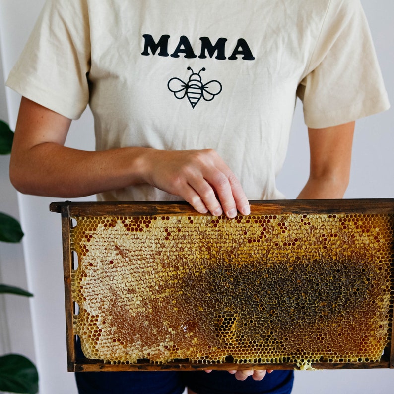K&ouml;nnte beinhalten: Eine Person h&auml;lt einen Holzrahmen mit einer Honigwabe darin. Der Rahmen ist mit goldener Honigwabe gef&uuml;llt. Die Person tr&auml;gt ein hellbeiges T-Shirt mit dem Wort "MAMA" darauf und einer schwarzen Bienenillustration.