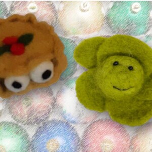 Puede incluir: Dos adornos de fieltro caprichosos. Uno es un pastel con ojos saltones, bayas de acebo y una hoja verde. El otro es una criatura verde con forma de flor y una cara sonriente. El fondo es un patrón borroso de adornos navideños coloridos.