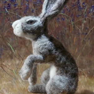 Puede incluir: Escultura de liebre hecha a mano en fieltro gris y blanco, posando sentada con una pata levantada. La liebre tiene orejas largas y detalles precisos. El fondo presenta un patrón floral borroso. Una pieza única.