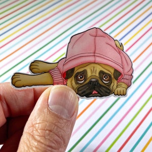 May include: A sticker of a pug wearing a pink hoodie. The pug is lying down with its head resting on its paws. The sticker is on a white and colorful striped background.