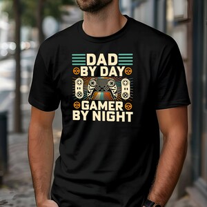 May include: Black t-shirt featuring a retro-style graphic that reads "Dad by Day Gamer by Night" with a stylised video game controller in the centre.