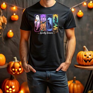 May include: Black t-shirt with a colorful Halloween-themed graphic. The design features a ghost, pumpkin, black cat, and haunted house. The words "Spooky Season" are printed below the images. The background includes pumpkins and decorative lights.