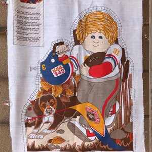 May include: A Cabbage Patch Kids sewing pattern featuring a football player with a dog. The pattern is printed on white fabric with brown, red, blue, yellow, and white details. The pattern includes instructions for sewing the doll and dog. The text on the pattern reads "Cabbage Patch Kids Collection Pattern "Down the Line" 1983 Original Appalachian Arts & Crafts, Inc."