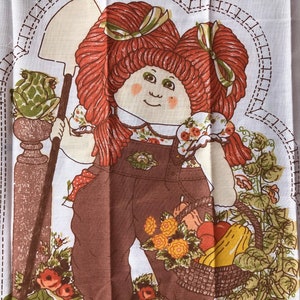 May include: A vintage fabric panel featuring a cartoon character with red pigtails, wearing brown overalls and holding a shovel. The character is surrounded by a garden scene with vegetables, flowers, and a basket of produce. The panel has a white background.
