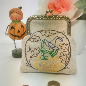 May include: A small, beige linen coin purse with a metal clasp. The purse is decorated with an embroidered design of a cat wearing a witch's hat and sitting inside a pumpkin. The design is surrounded by leaves.