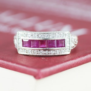 May include: A silver band ring with a row of four square-shaped pink rubies surrounded by smaller diamonds.