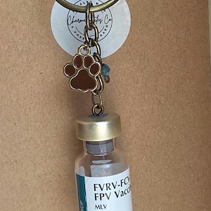 May include: A bronze-toned keychain featuring a small brown paw print charm and a miniature glass vial labeled FVRV-FCV FPV Vaccine MLV VANGUARD Feline RCP Injectable. The keychain includes a circular ring and a small blue bead accent.