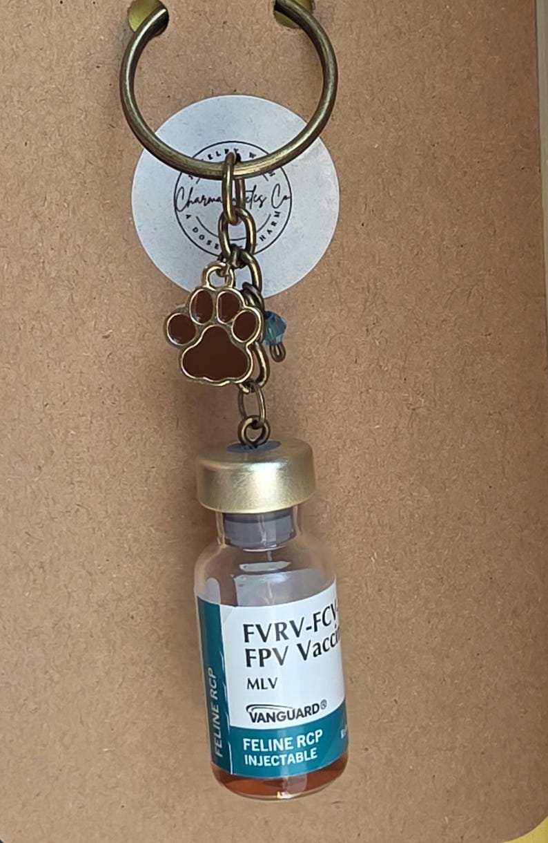 May include: A bronze-toned keychain featuring a small brown paw print charm and a miniature glass vial labeled FVRV-FCV FPV Vaccine MLV VANGUARD Feline RCP Injectable. The keychain includes a circular ring and a small blue bead accent.