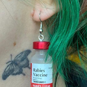 May include: A pair of earrings shaped like miniature vials of rabies vaccine. The vials are red and clear with a white label that reads "Rabies Vaccine, Killed Virus, VANGUARD, Rabies 1 Year Injectable".