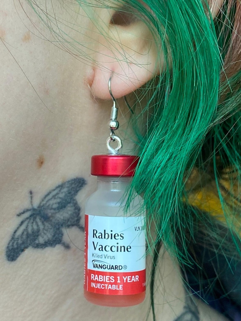 May include: A pair of earrings shaped like miniature vials of rabies vaccine. The vials are red and clear with a white label that reads "Rabies Vaccine, Killed Virus, VANGUARD, Rabies 1 Year Injectable".