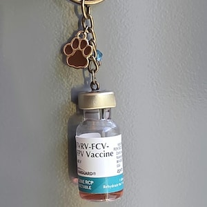 May include: A keychain featuring a bronze-toned metal ring and chain, a small brown paw print charm, a tiny blue bead, and a miniature glass vial labeled FVRV-FCV-FPV Vaccine with a gold-colored cap and a teal band at the bottom of the label.