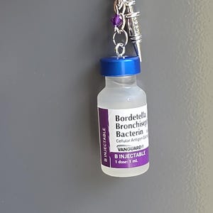 May include: A keychain featuring a miniature clear glass vial with a blue cap and a purple label reading Bordetella Bronchiseptica Bacterin, Vanguard, and B Injectable. A small silver syringe charm and a purple bead are attached to the metal ring.