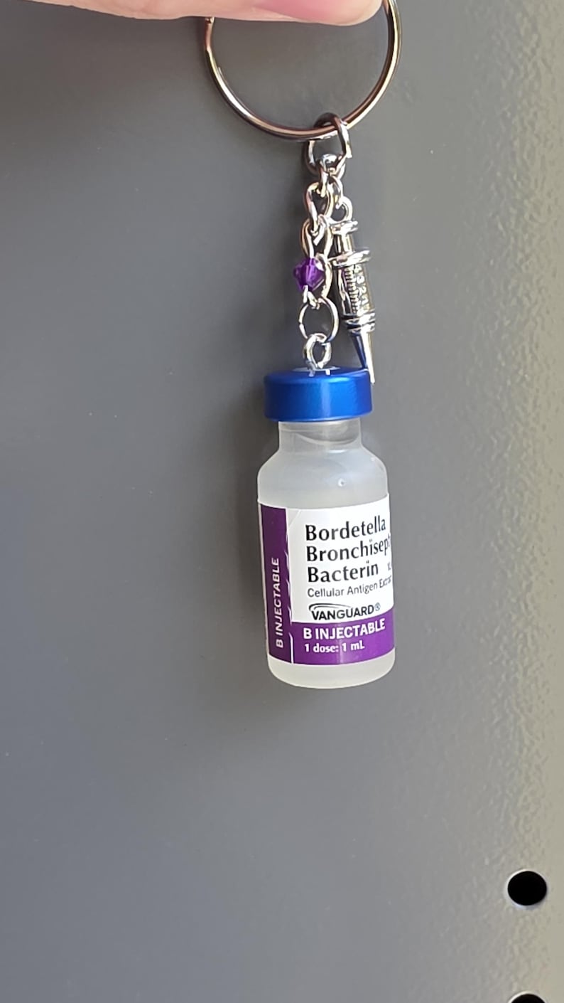 May include: A keychain featuring a miniature clear glass vial with a blue cap and a purple label reading Bordetella Bronchiseptica Bacterin, Vanguard, and B Injectable. A small silver syringe charm and a purple bead are attached to the metal ring.
