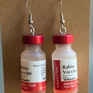 Puede incluir: Un par de pendientes de vial de vidrio rojo y transparente con ganchos plateados. Los viales están etiquetados como "Rabies Vaccine" y "Babies Vaccine" con el texto "Rabies 1 Year Injectable" y "Babies 1 Year Injectable".
