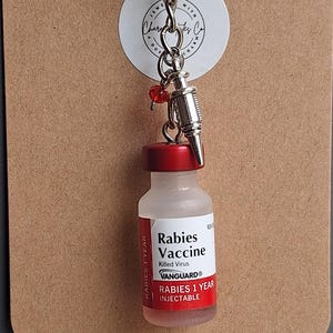 May include: A keychain featuring a miniature Rabies Vaccine vial with a red cap and a label reading Rabies Vaccine Killed Virus VANGUARD RABIES 1 YEAR INJECTABLE. It includes a small silver syringe charm and a single red bead attached to a metal ring.