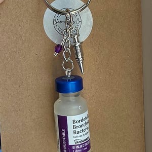 May include: A keychain featuring a miniature glass vial with a blue cap and a label reading Bordetella Bronchiseptica Bacterin, Vanguard B Injectable, 1 dose: 1 ml. Attached to the silver ring are a small silver syringe charm and a purple bead.