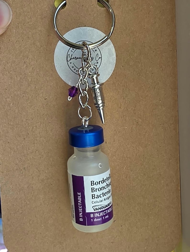 May include: A keychain featuring a miniature glass vial with a blue cap and a label reading Bordetella Bronchiseptica Bacterin, Vanguard B Injectable, 1 dose: 1 ml. Attached to the silver ring are a small silver syringe charm and a purple bead.
