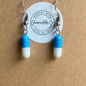 May include: A pair of novelty earrings featuring a blue and white capsule design. The earrings have silver-colored hooks and are displayed against a brown background. The earrings are from Charmacuticles Co.