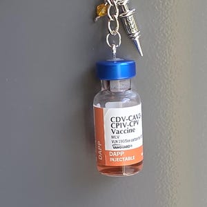 May include: A keychain featuring a miniature clear glass vial with a blue cap and an orange label reading CDV-CAV2 CPIV-CPV Vaccine DAPP INJECTABLE. Attached to the silver chain are a small silver syringe charm and a single yellow crystal bead.
