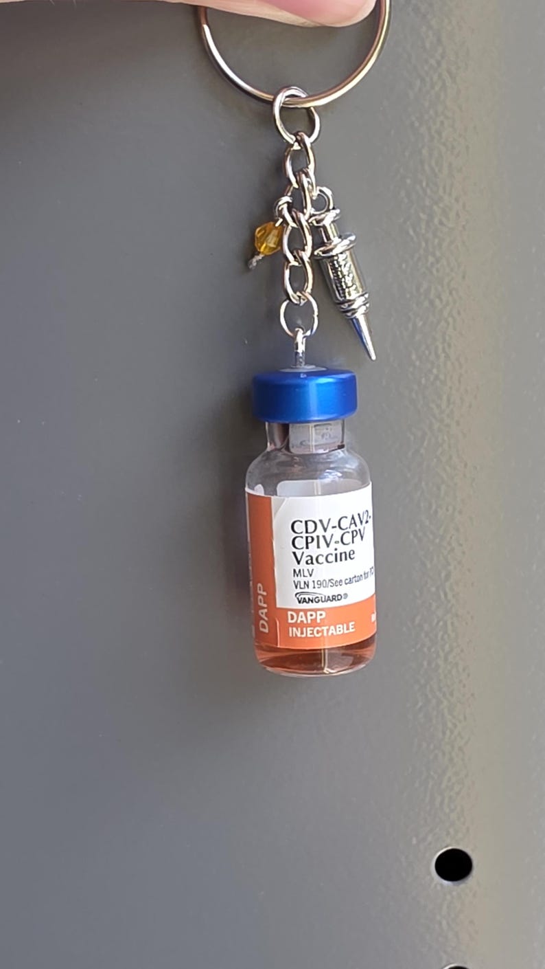 May include: A keychain featuring a miniature clear glass vial with a blue cap and an orange label reading CDV-CAV2 CPIV-CPV Vaccine DAPP INJECTABLE. Attached to the silver chain are a small silver syringe charm and a single yellow crystal bead.