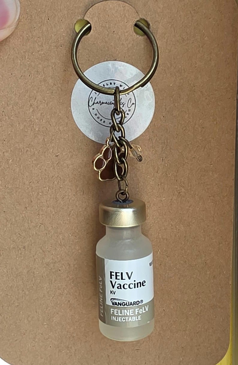 May include: A keychain featuring a bronze-toned metal ring and chain, a small brown paw print charm, and a miniature glass vial labeled FELV Vaccine KV Vanguard Feline FeLV Injectable. The keychain is attached to a brown cardboard backing card.