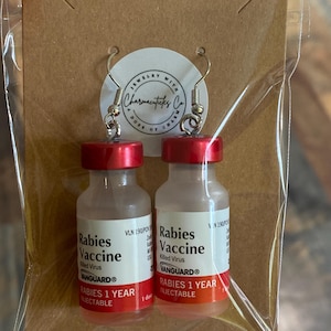 May include: A pair of earrings shaped like vials of rabies vaccine. The vials are clear glass with red tops and labels that read "Rabies Vaccine" and "VANGUARD&reg;". The labels also state "Rabies 1 Year Injectable".