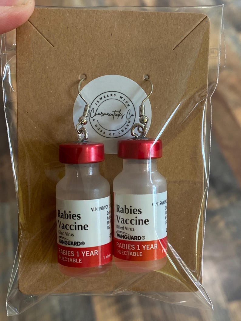 May include: A pair of earrings shaped like vials of rabies vaccine. The vials are clear glass with red tops and labels that read "Rabies Vaccine" and "VANGUARD&reg;". The labels also state "Rabies 1 Year Injectable".
