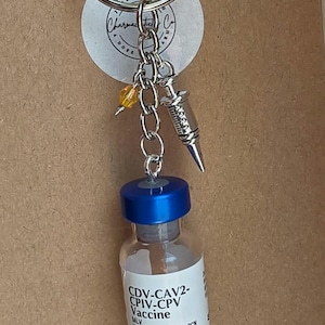 May include: A keychain featuring a silver metal ring, a small silver syringe charm, a single yellow bead, and a miniature glass vial with a blue cap. The vial label reads CDV-CAV2-CPIV-CPV Vaccine, MLV, VANGUARD, DAPP, and INJECTABLE in black and orange text.